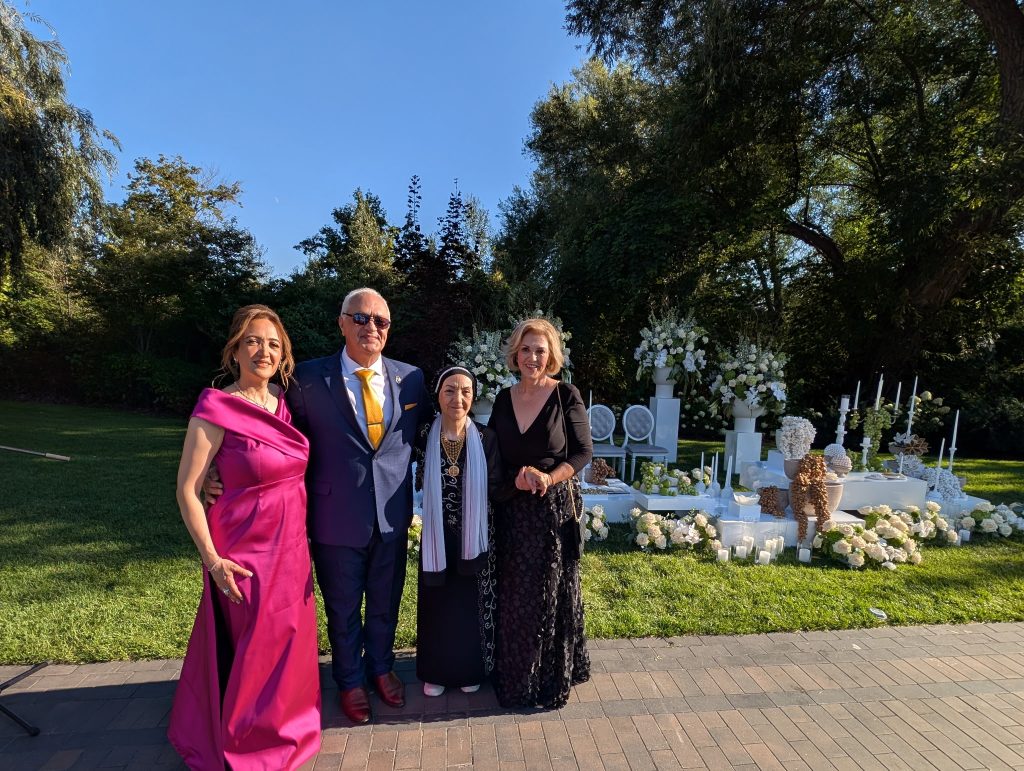 Daryoush Ghashghai, picture with bride's family 1 of 2 Daryoush Ghashghai officiating a bilingual wedding ceremony in Canada, standing with the bride’s family beside a traditional sofreh aghd setup in an outdoor garden (image 1 of 2)
